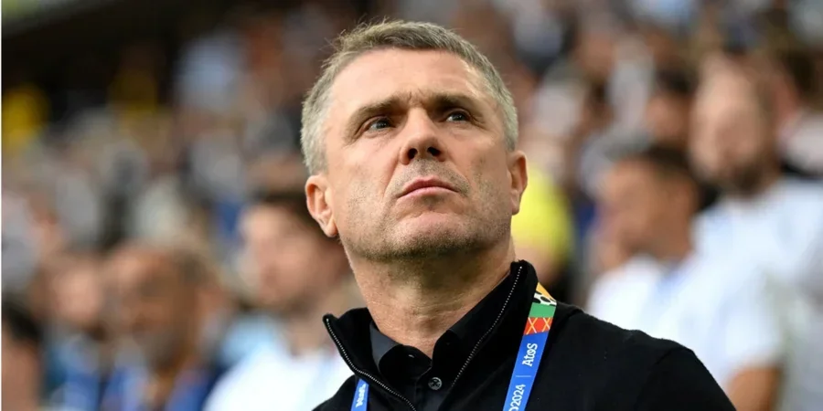 Сергій Ребров у чорній куртці з бейджем дивиться вгору на стадіоні