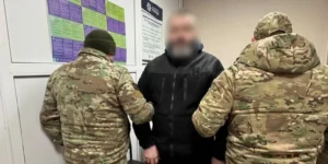 Двоє військових у камуфляжі утримують чоловіка в чорній куртці з розмитим обличчям
