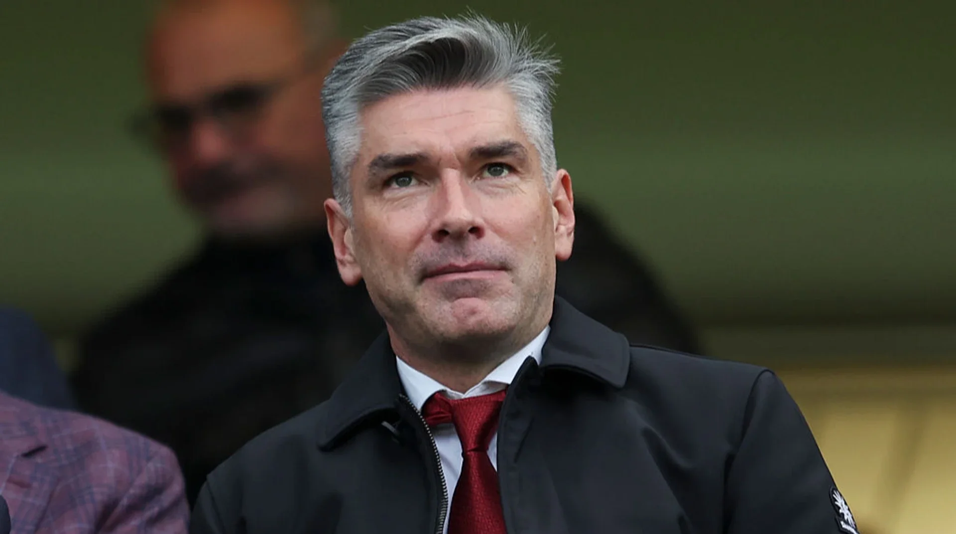 Сіроволосий чоловік у чорній куртці та червоній краватці на трибуні