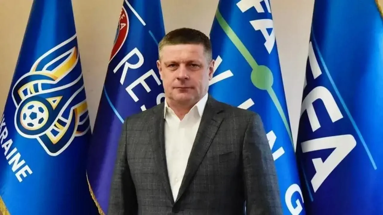 Чоловік у сірому костюмі стоїть перед прапорами Української асоціації футболу